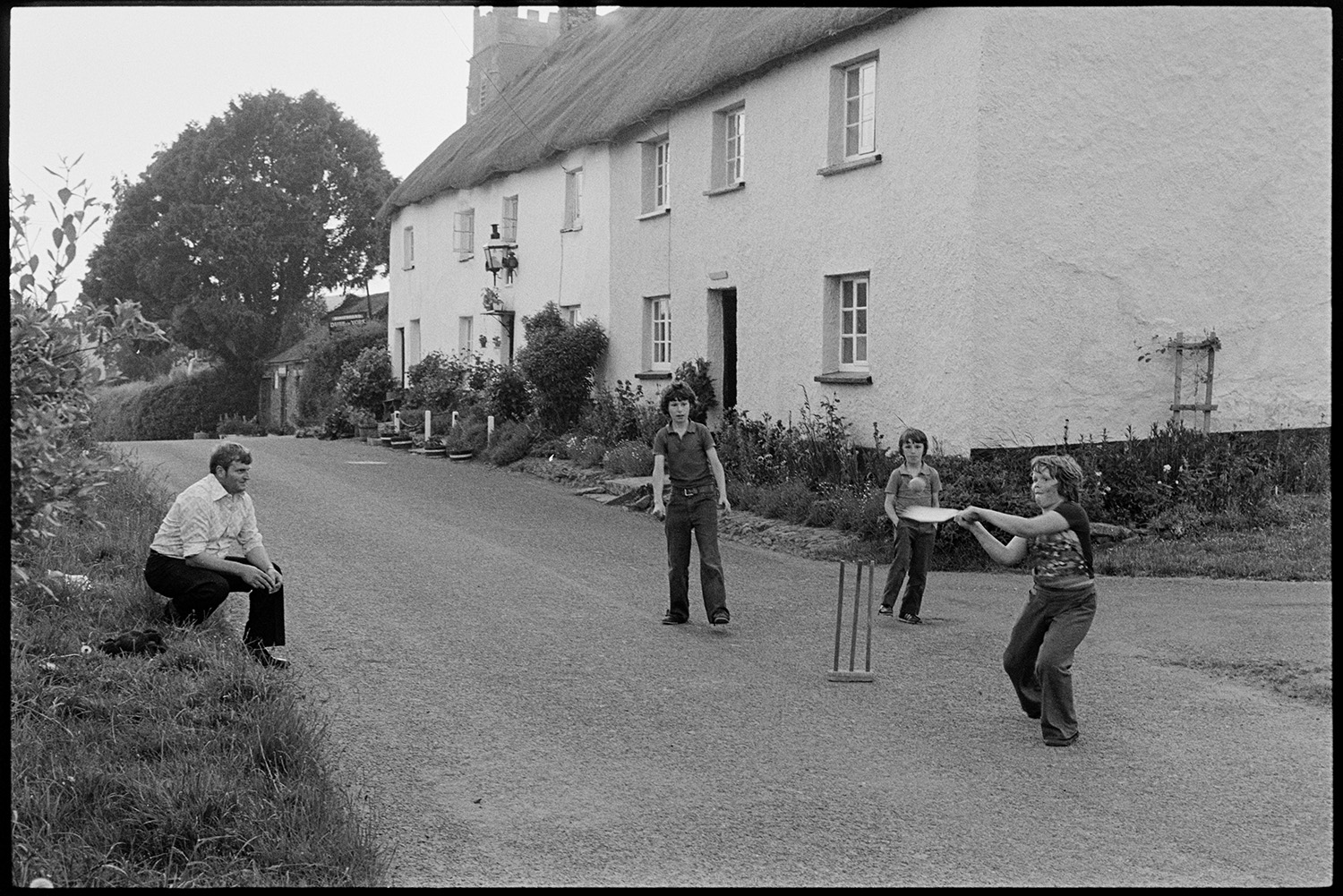 Children playing cricket in street. 
[A man watching three boys playing cricket in a street in Iddesleigh. One boy is about to hit the ball. Thatched cottages are visible in the background.]