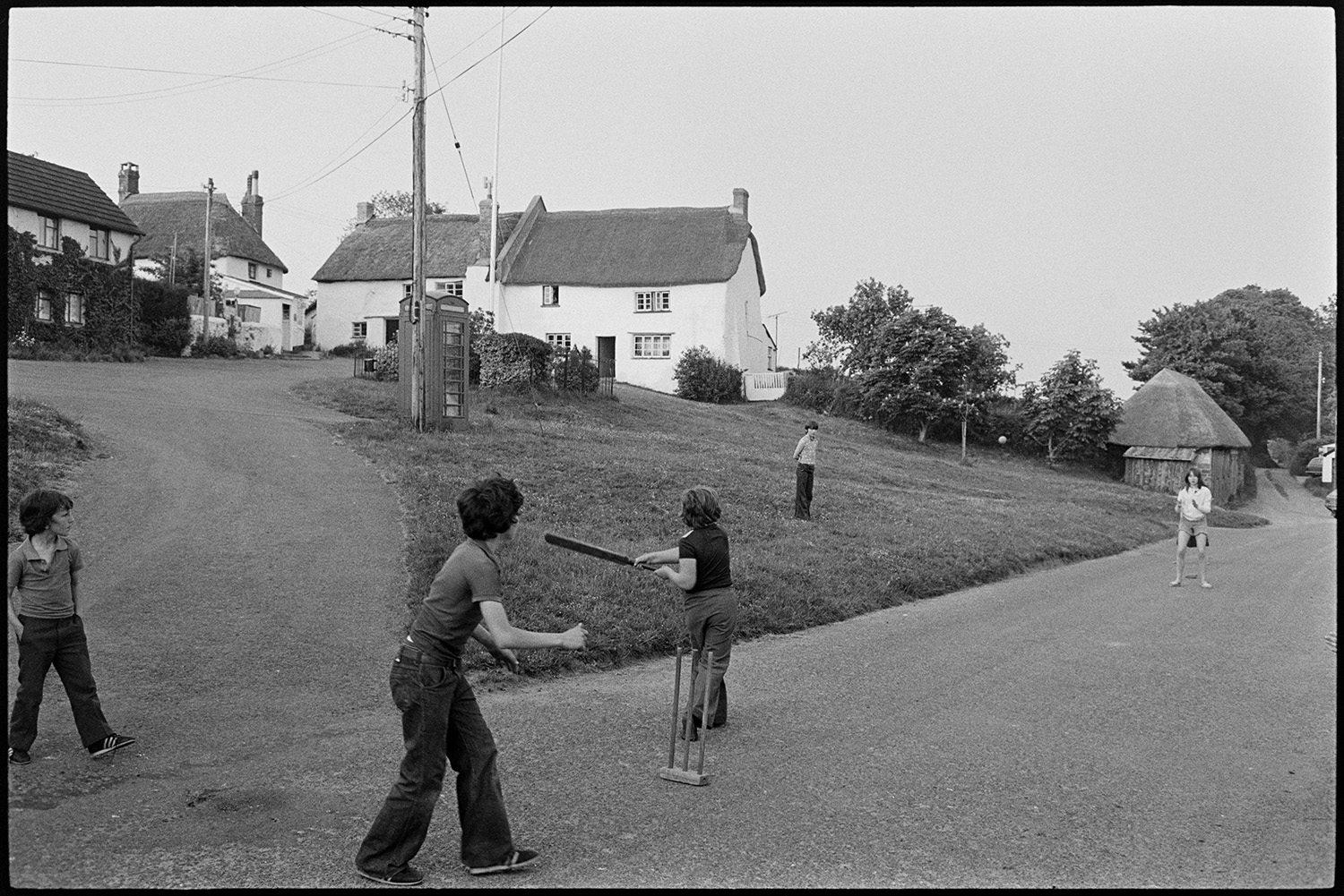 Children playing cricket in street. 
[Boys playing cricket in a street in Iddesleigh. Thatched cottages and a telephone box can be seen by the village green.]
