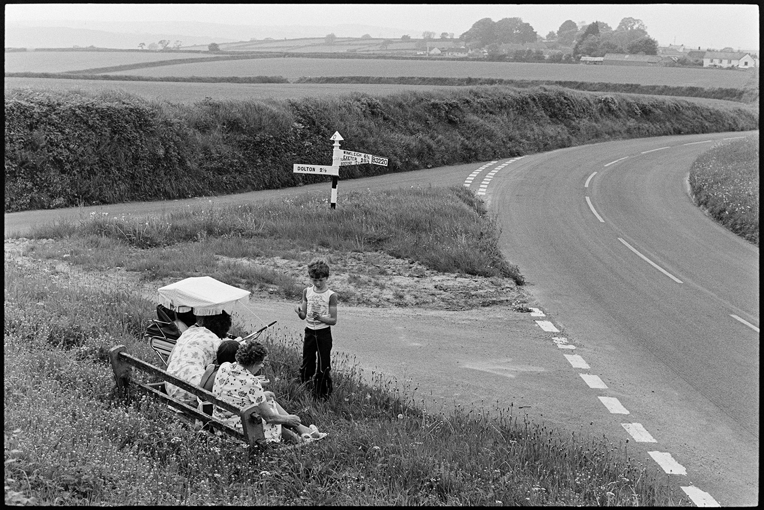 Family sitting beside road junction, now altered. 
[Two women and two children sitting on a wooden bench by a roadside junction at Beaford. They also have a pram with them. A signpost is by the road junction and fields can be seen in the background.]