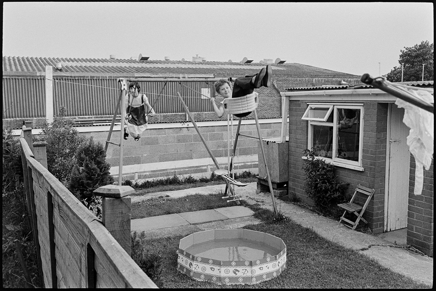 Children playing on swings. 
[Two children playing on swings in a garden in Dolton. A paddling pool is in front of the swings.]