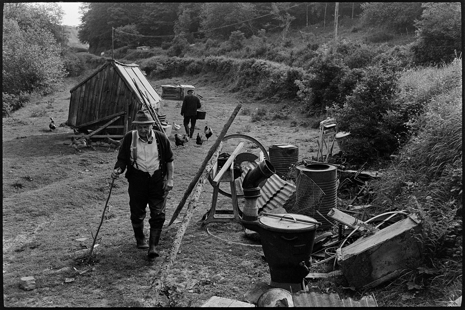 Two farmers sitting in field chatting, chickens and poultry house. Straw cutter. 
[Archie Parkhouse walking past an assortment of items, including barrels, a straw cutter and piping, in a field at Millhams, Dolton. He is smoking a pipe. In the background Ivor Brock is carrying a bucket past a poultry house and chickens, possibly feeding them.]