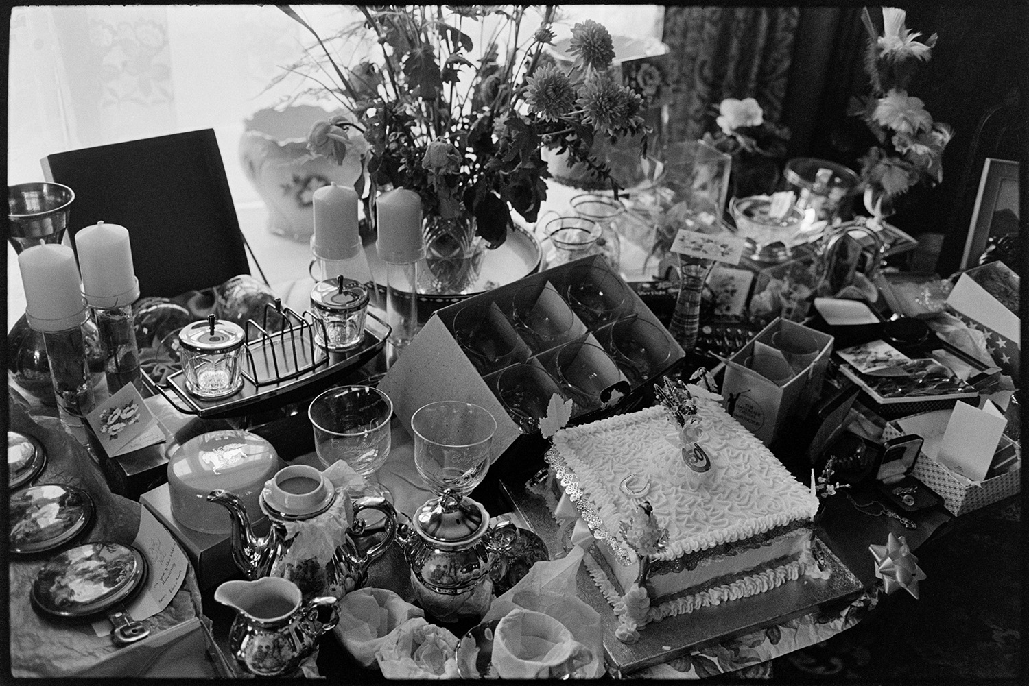 Golden Wedding Anniversary presents, cake and flowers. 
[Presents given to Mr and Mrs Hutchins for their Golden Wedding anniversary, displayed in their home at Fore Street, Dolton. Gifts includes teapots , glassware and candles. A cake is in the middle of the presents.]