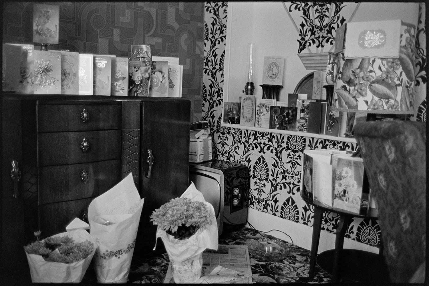 Golden Wedding Anniversary presents, cake and flowers. 
[Cards and flowers given to Mr and Mrs Hutchins for their Golden Wedding anniversary, displayed in their home at Fore Street, Dolton. The walls are covered with patterned wallpaper.]