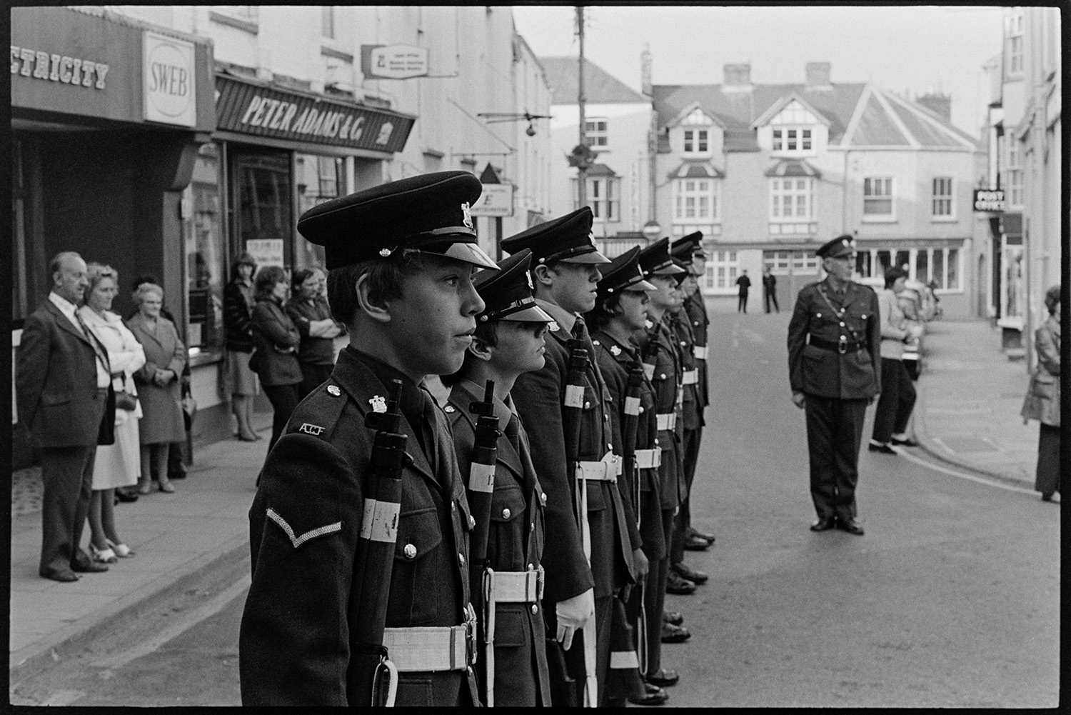 Military parade round town with Brass Band. 
[Boys lined up in uniform and holding guns, for a military parade in Torrington. Spectators are watching from the side of the street, outside shop fronts.]