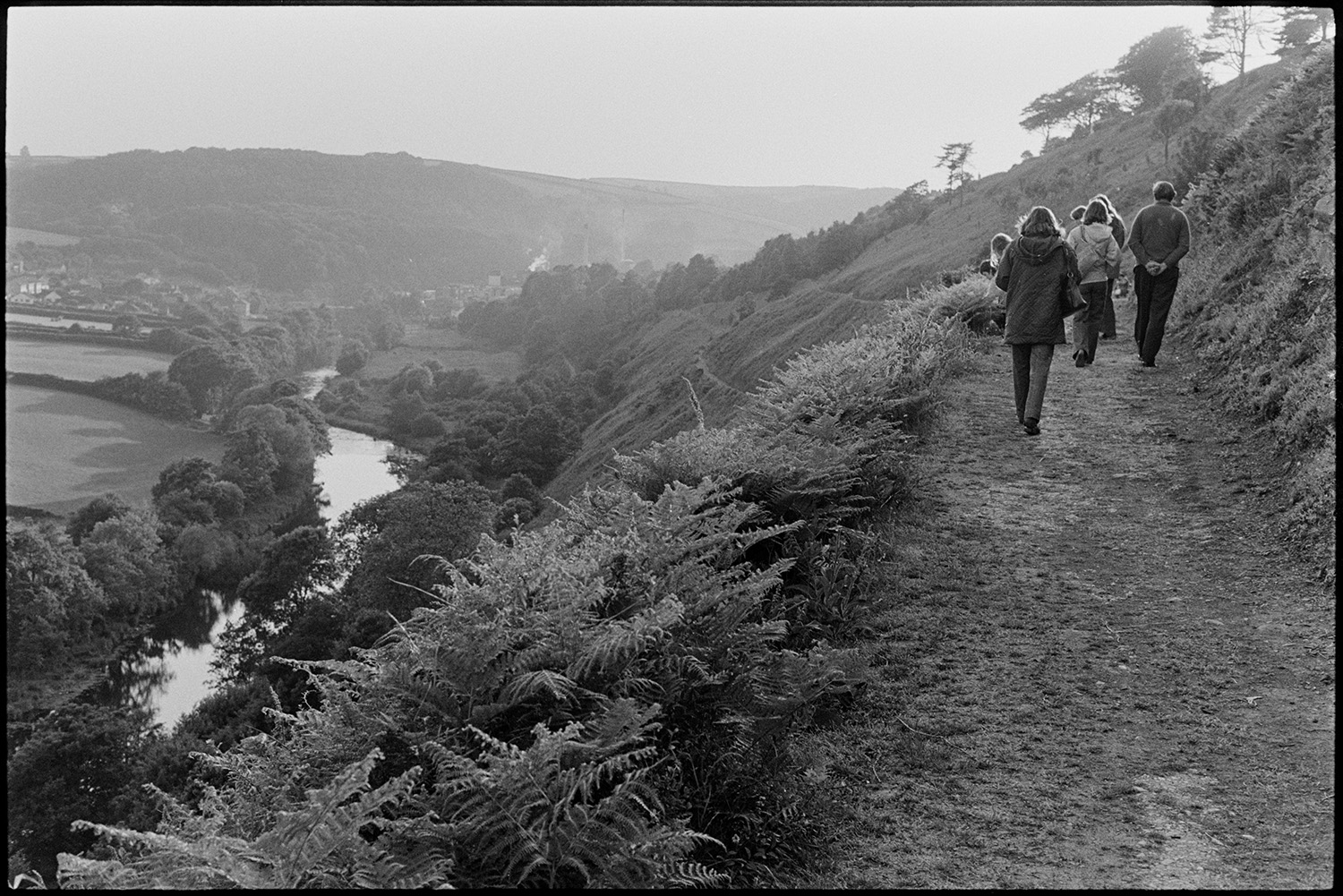 Landscape with river and bales waiting to be collected from distant fields, walkers on path. 
[People walking along a path, possibly at Castle Hill Park, in Torrington. The River Torridge can be seen in the valley below, running past fields.]