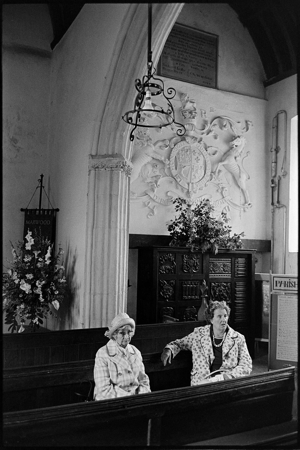Two women in church looking at flower display. 
[Two women sat in the pews in Marwood Church looking at the Flower Show in the church. Two flower displays can be seen in the background.]