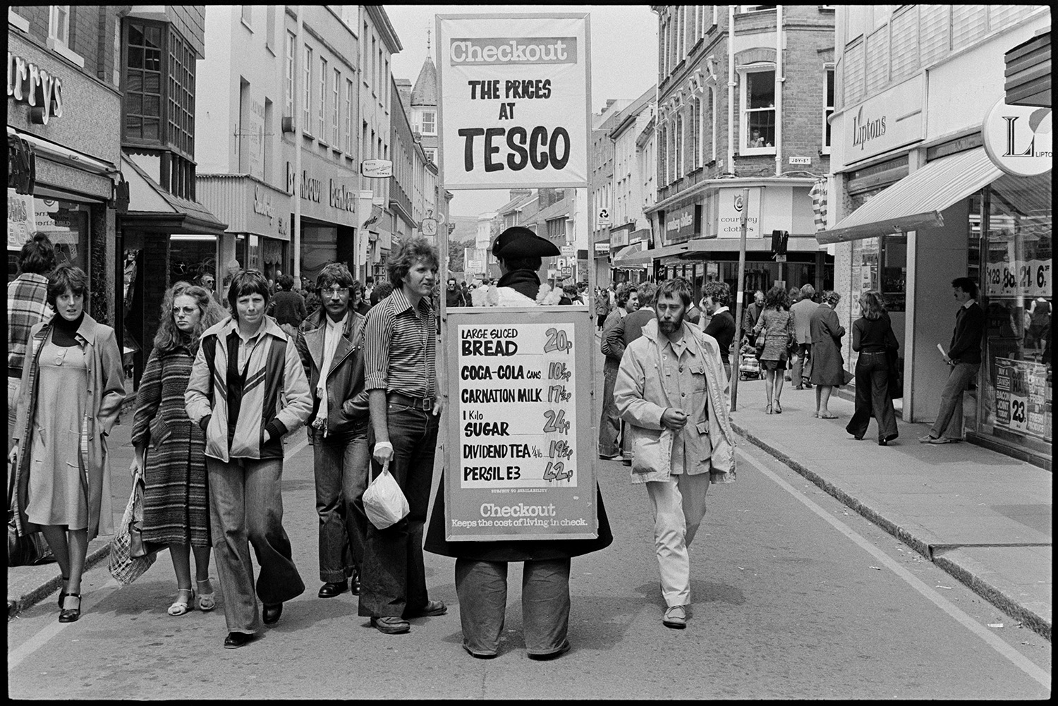 Sandwich board man in town street. 
[A man wearing a sandwich board and tricorn hat advertising Tesco in Barnstaple High Street. Shoppers are walking along the street and various shop fronts are visible, including Liptons and Bembows.]