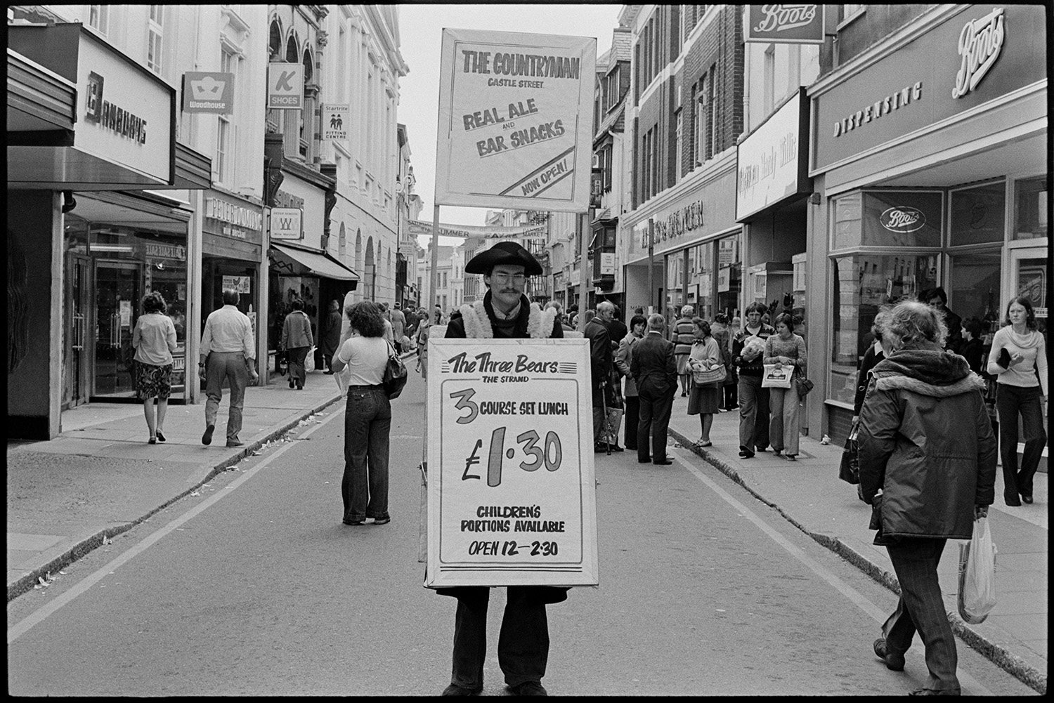 Sandwich board man in town street. 
[A man wearing a sandwich board and tricorn hat advertising The Countryman and The Three Bears pubs in Barnstaple High Street. Shoppers are walking along the street and various shop fronts are visible, including Boots and Marks & Spencer.]