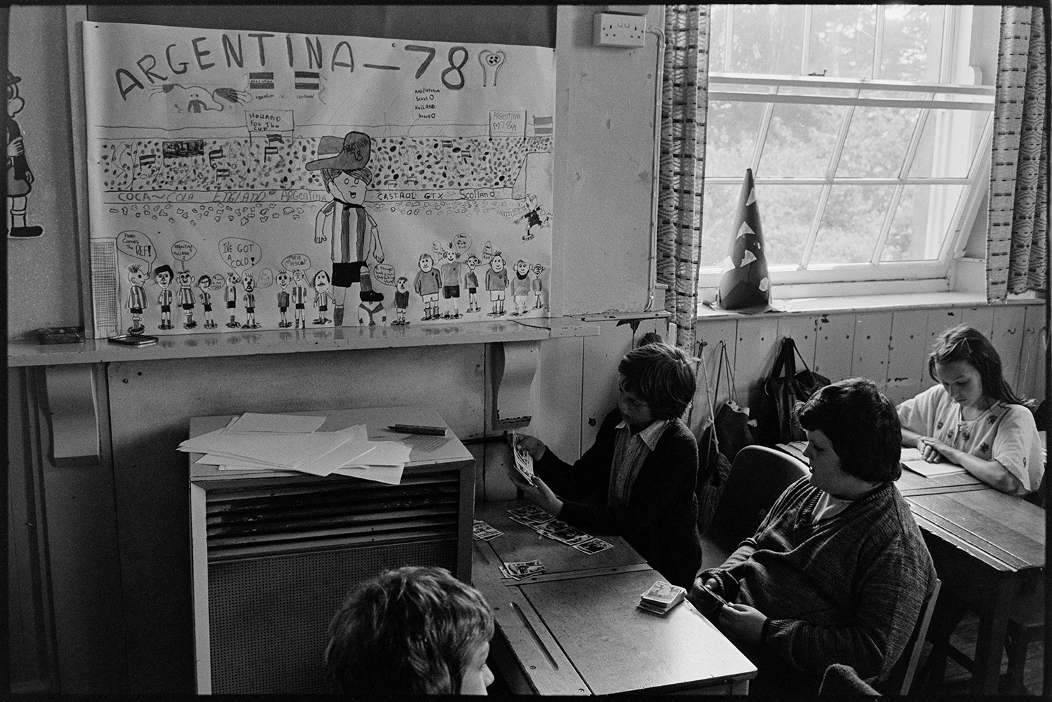 Children in class, arranging flowers. 
[Children in a classroom at Blue Coats School, Torrington. Two of them are looking at cards and a girl in the background is reading. A poster on the wall shows a child's drawing of the FIFA World Cup in Argentina in 1978.]