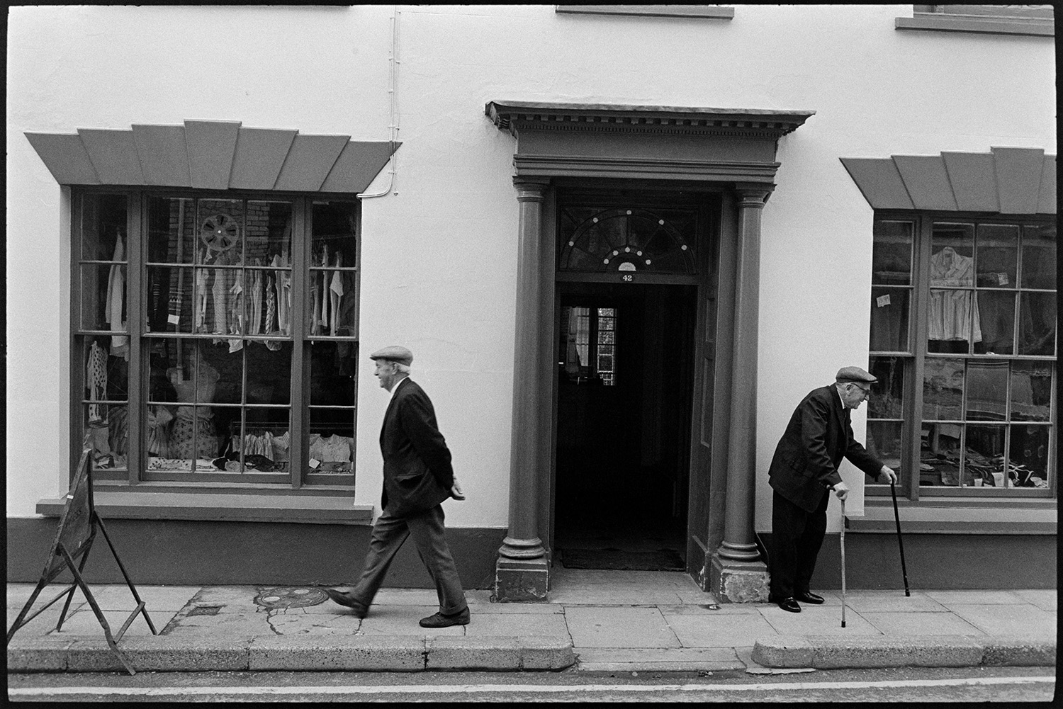Two old men chatting in front of fine house, built by William Corey. 
[Two men passing in the street outside a house built by William Corey in South Street, Torrington. The house is now a clothes shop and clothes are displayed in the windows. One of the men is using two walking sticks.]