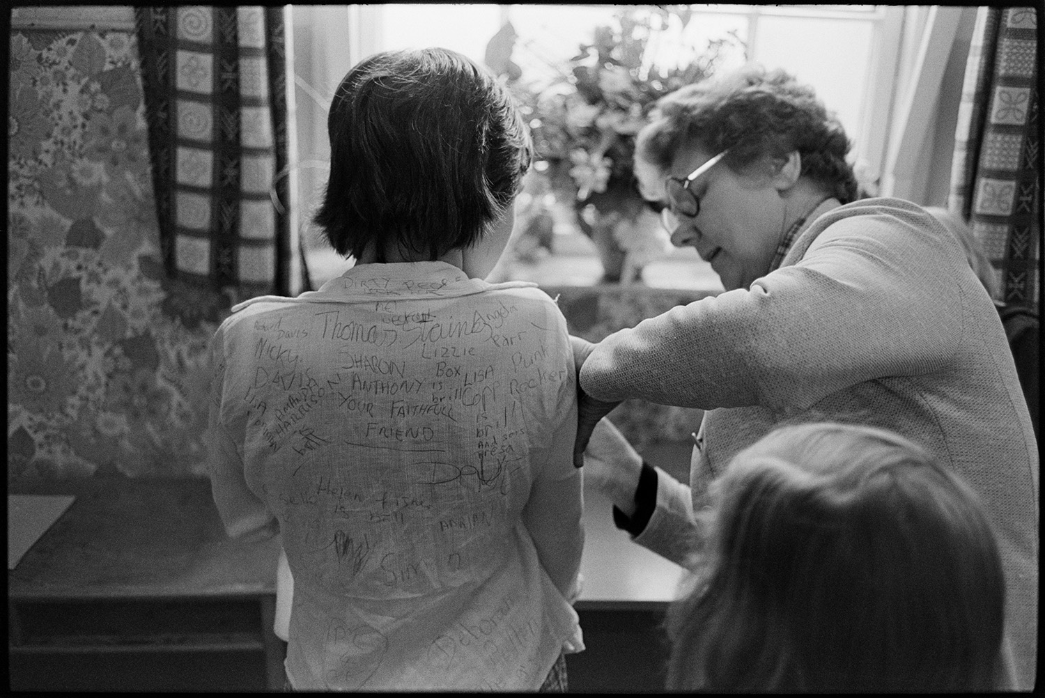 Children arriving at school and getting ready for assembly. 
[A teacher signing a child's shirt at Blue Coats School, Torrington. Other signatures and messages can be seen on the back of the child's shirt.]