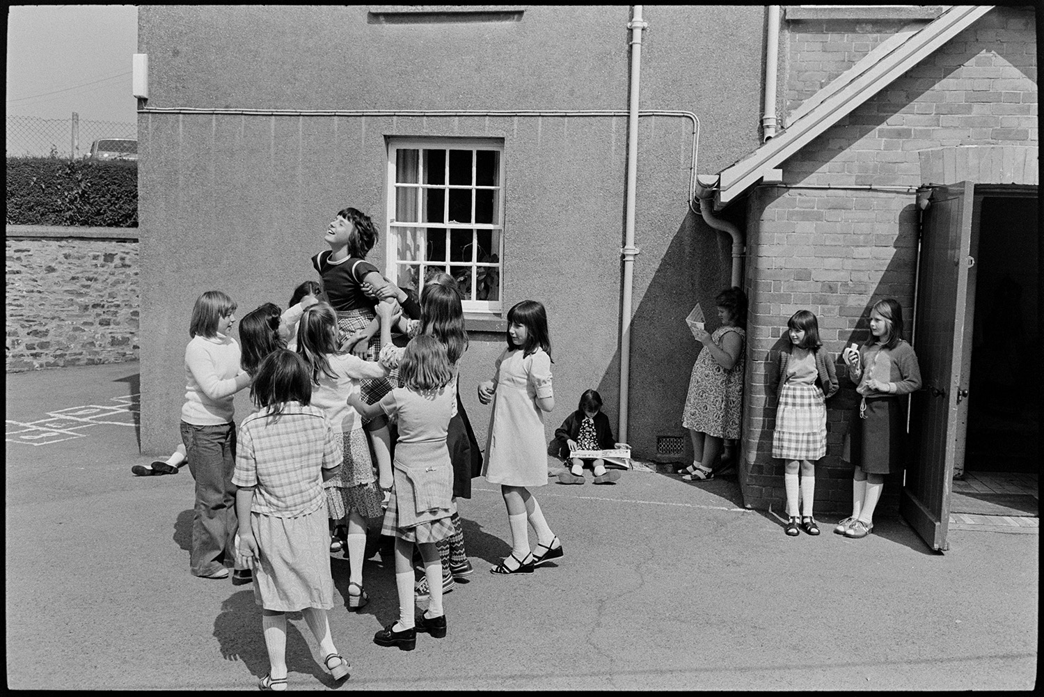Children at break time, having free milk and playing outside. 
[Children playing in the playground at Blue Coats School in Torrington at break time. They are lifting up one of their classmates. In the background one child is reading and another is eating a banana.]