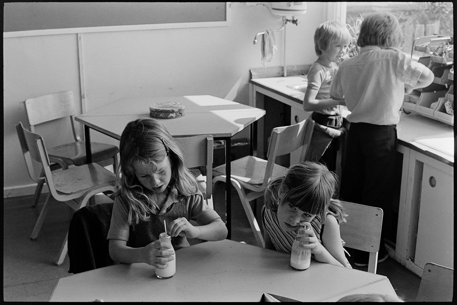 Children at break time, having free milk and playing outside. 
[Two girls drinking their free milk in a classroom at Blue Coats School in Torrington. Other children are in the background.]
