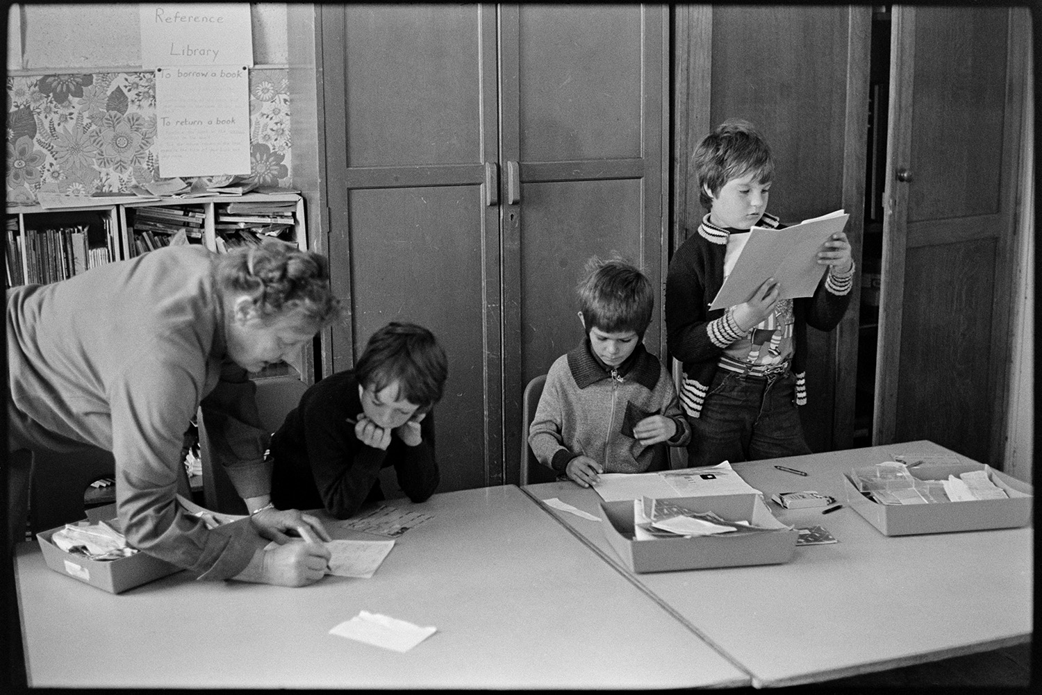 Schoolchildren and teachers in class, reading and asking questions. 
[Boys working at desks in a classroom at Blue Coats School in Torrington. A teacher is checking one of the boys' work. A bookcase is visible in the background.]