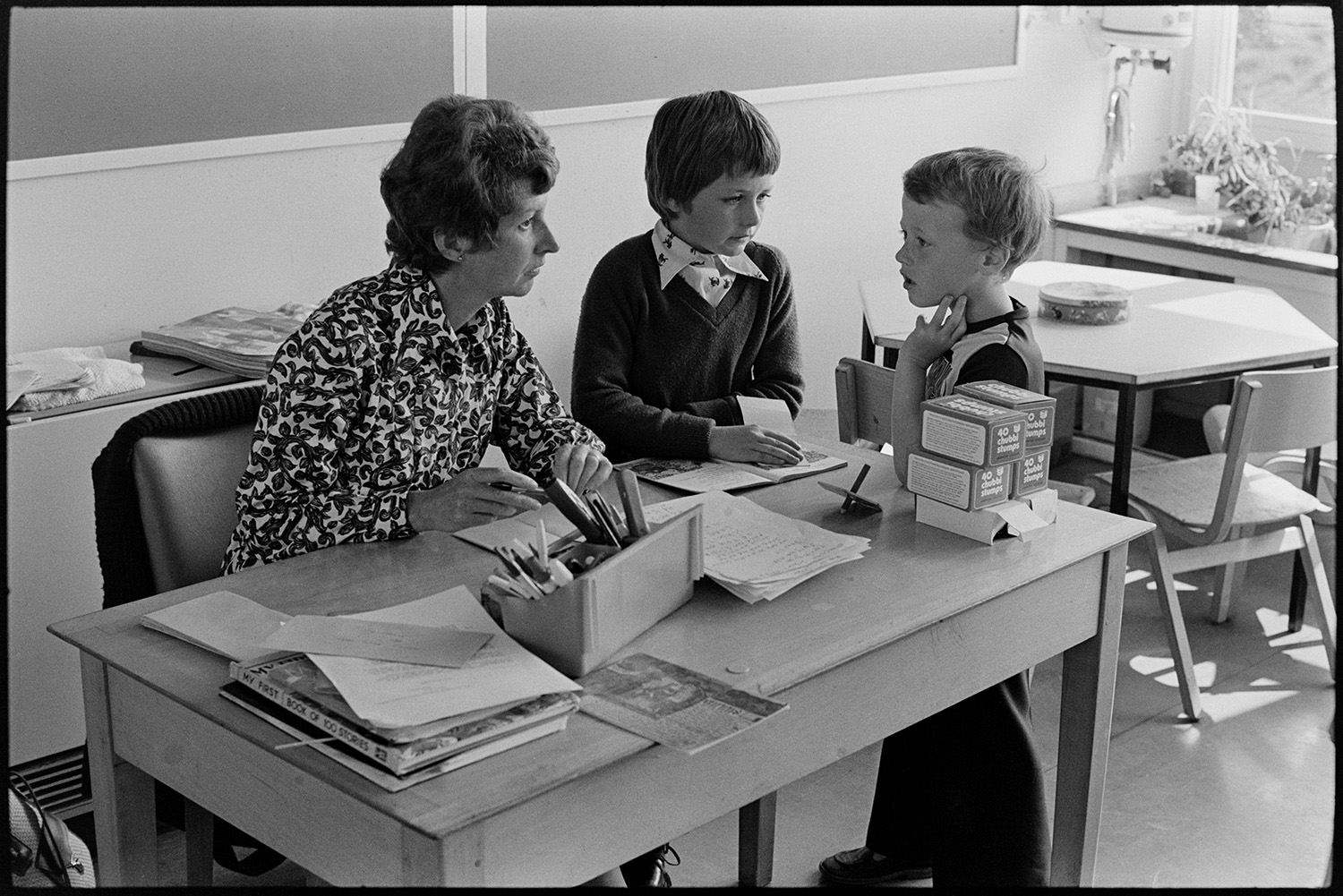 Schoolchildren and teachers in class, reading and asking questions. 
[A teacher sat at her desk in a classroom in Blue Coats School in Torrington. She is talking to two boys. One of the boys is reading a book to her.]