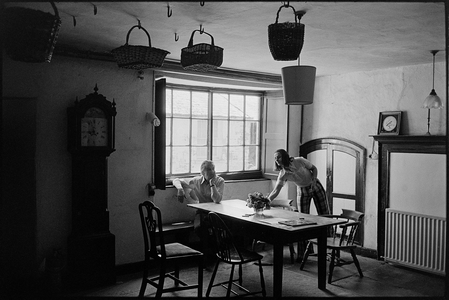 People sitting round kitchen table of large country house. 
[Pat Furse and Antonia Furse sat and stood by the kitchen table in Halsdon House in Dolton. A grandfather clock is positioned against a wall and baskets are hung from the ceiling.]