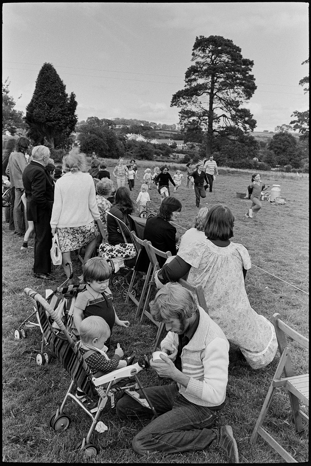 Flower show with sports and display of produce. 
[Spectators, including men, women and children, watching a children's running race in a field, at Dolton Flower Show.  In the foreground an man is feeding a baby,]