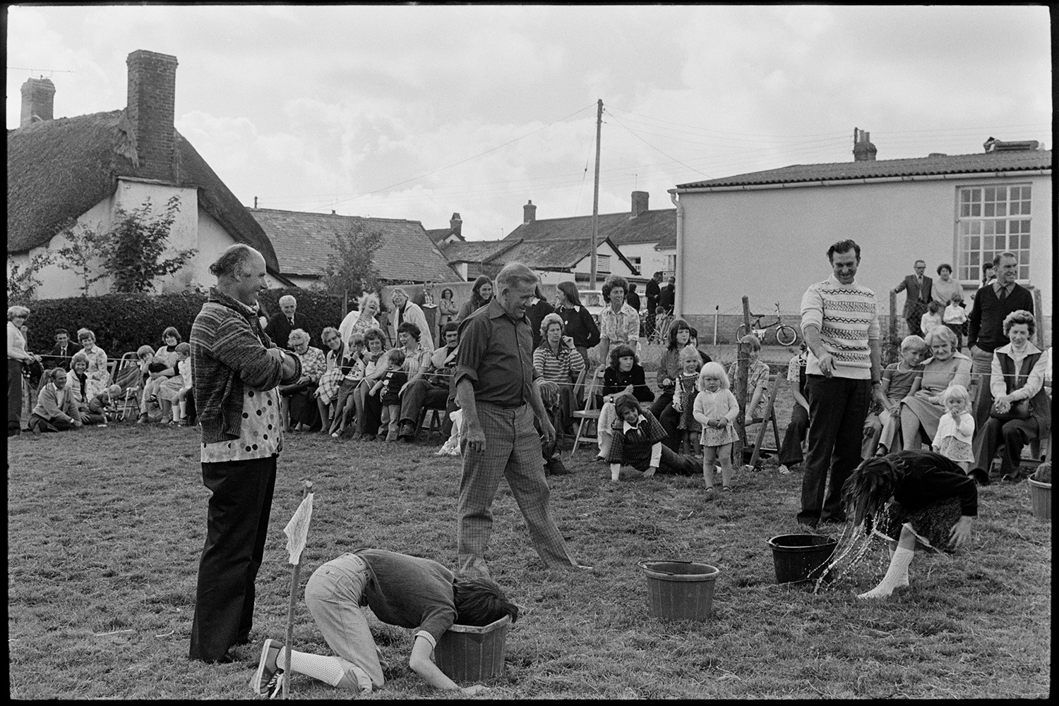 Flower show with sports and display of produce. 
[Spectators, including men, women and children, watching a children's sports event with buckets of water, at Dolton Flower Show.]