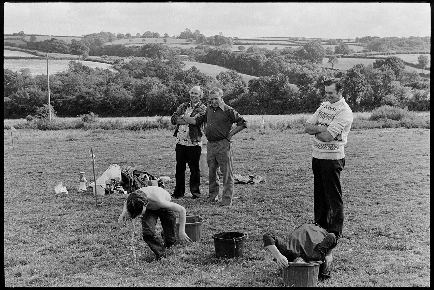 Flower show with sports and display of produce. 
[Children taking part in a sports even with buckets of water in a field, at Dolton Flower Show. Three men are supervising the sports.]