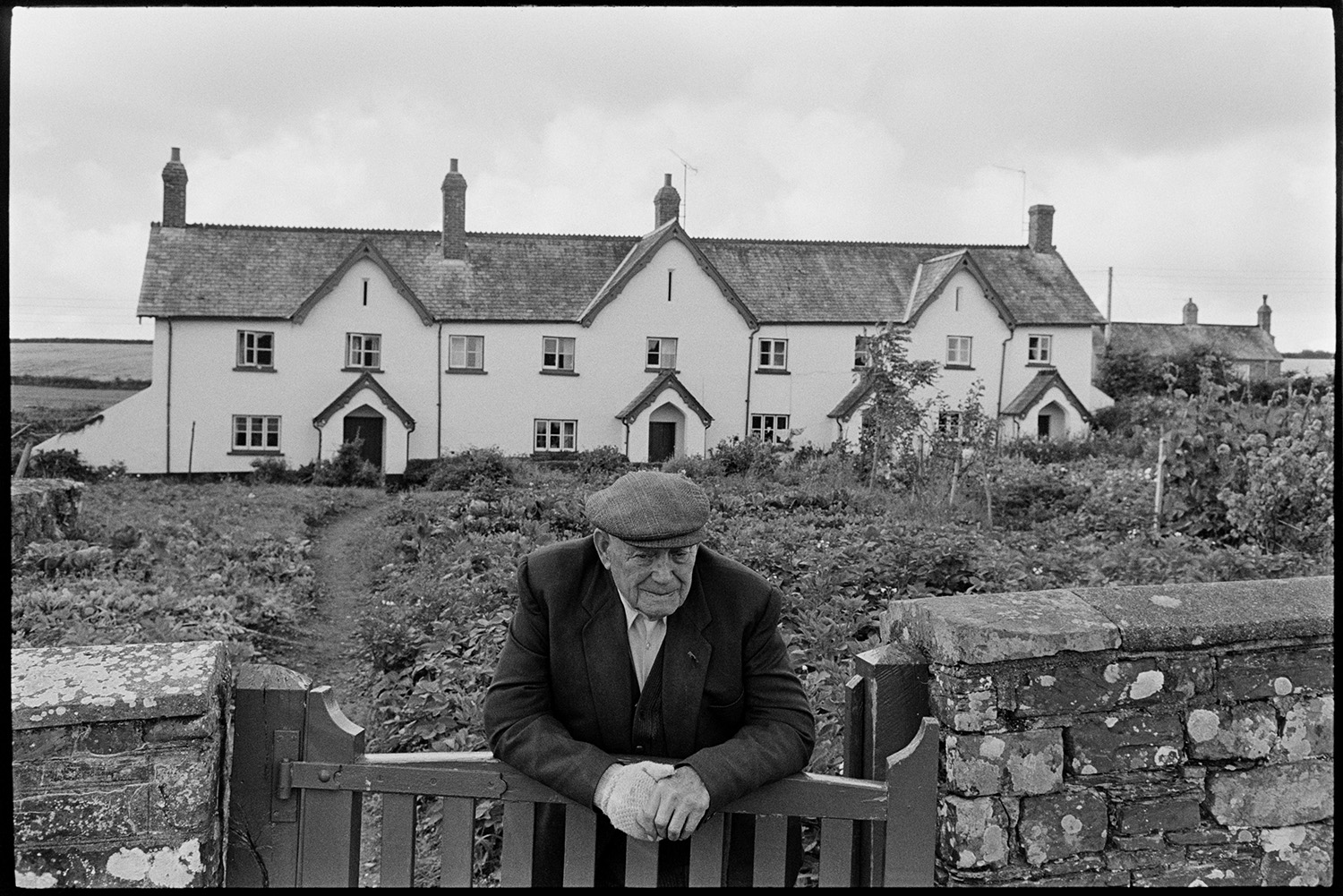 Man leaning over garden gate in front of estate cottages. 
[A man leaning on the gate at the entrance to terraced estate cottages and gardens at Merton.]