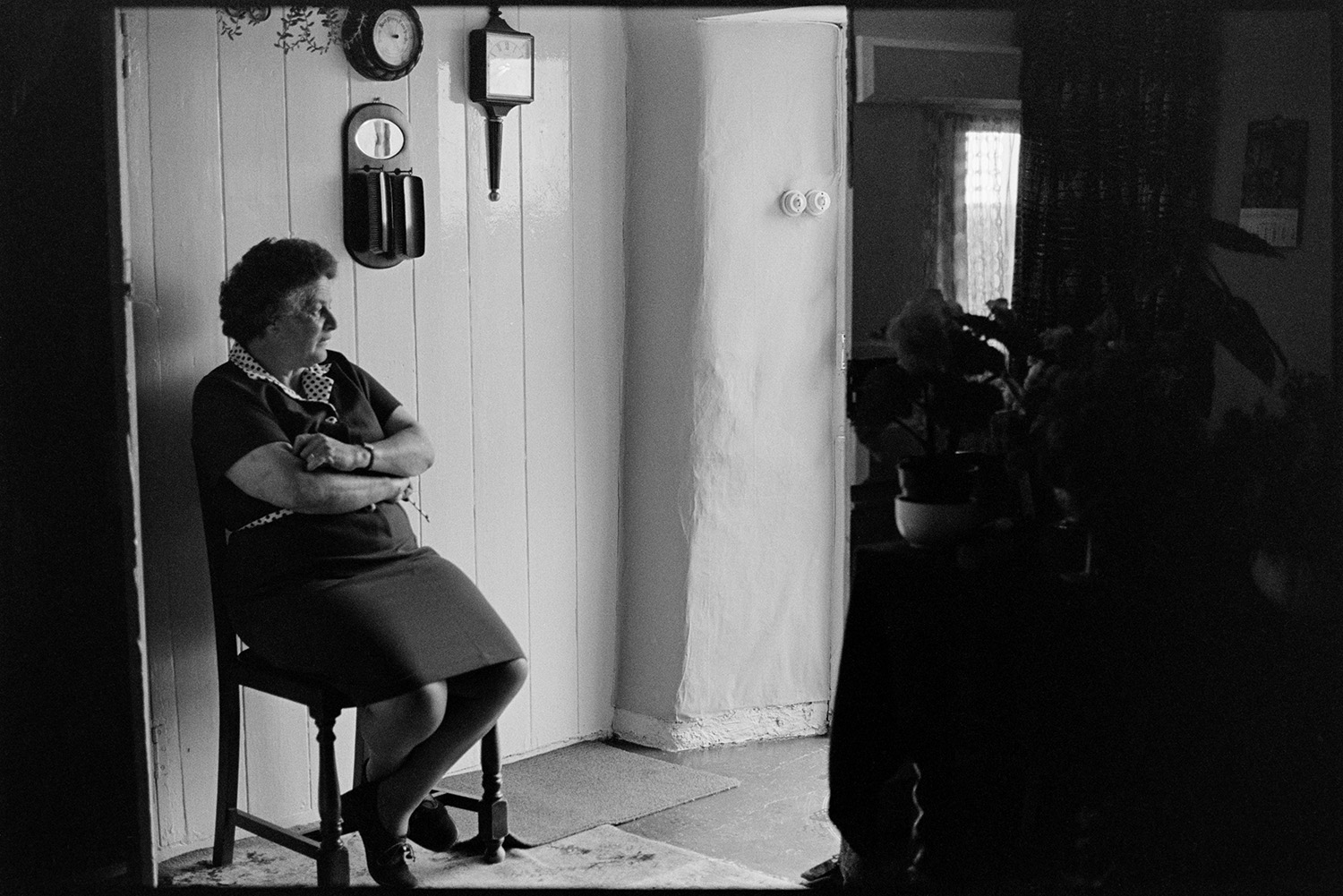Cottage interior, woman with flowers and sitting at doorway getting ready for show. 
[Mrs Piper sitting on a chair by the doorway to her cottage in Upcott, Dolton, waiting to go to Dolton Flower Show. A mirror and barometer are hung on the wall behind her.]
