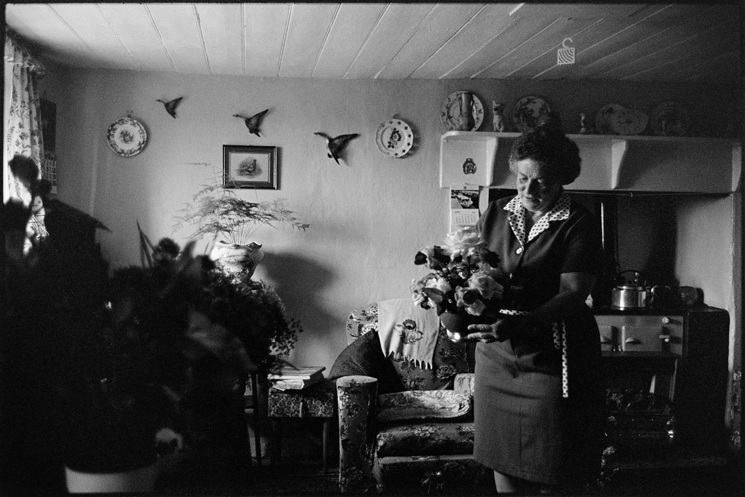 Cottage interior, woman with flowers and sitting at doorway getting ready for show. 
[Mrs Piper in her house at Upcott, Dolton. She is holding a vase of flowers, ready for Dolton Flower Show. An armchair, rayburn stove and mantelpiece with a display of china can be seen in the background. Ornamental china flying ducks are also decorating the wall.]