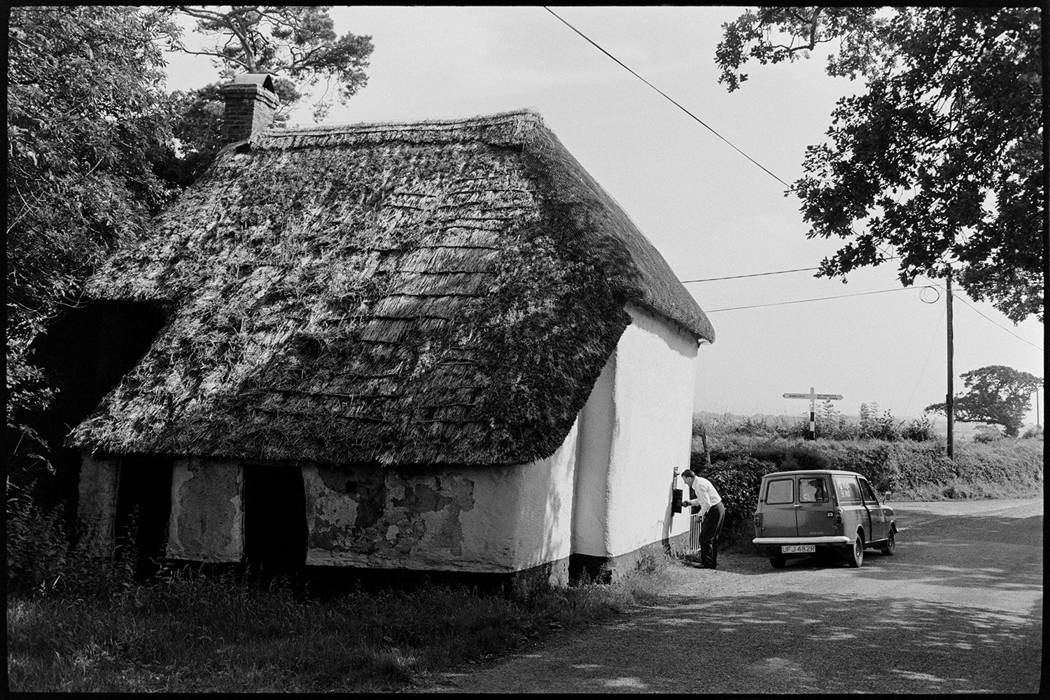 Sloping roof, thatched cottage from rear with postman emptying letter box, post van. 
[A postman collecting letters from a post box in the wall of the thatched cottage with a sloping roof at Nethercott Cross, Iddesleigh. His post van is parked on the road outside the cottage and a signpost can be seen in the hedge in the background.]