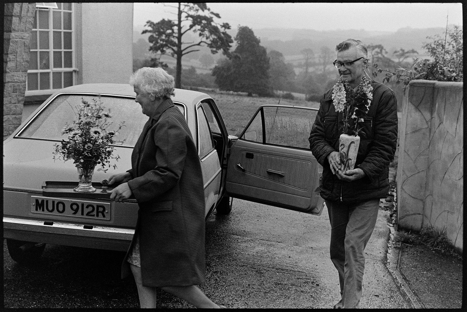 People bringing in entries for Flower show at village hall from cars. 
[A man and woman carrying vases of flowers from a parked car into Dolton Flower Show, at Dolton Village Hall.]