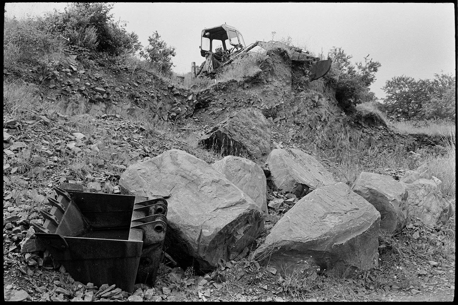 Abandoned machinery in quarry tractors, shovels. 
[A digger, digger bucket and boulders at Newbridge Quarry, Dolton.]