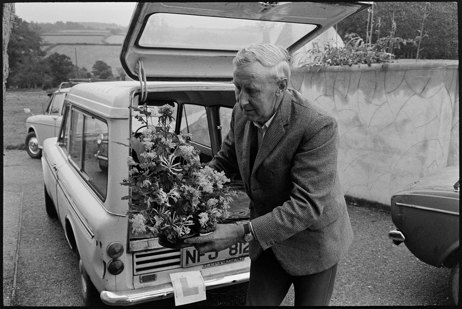 People bringing in entries for flower show and arranging them in village hall. 
[A man unloading a flower arrangement from a car and taking it into Dolton Village Hall for Dolton Flower Show.]