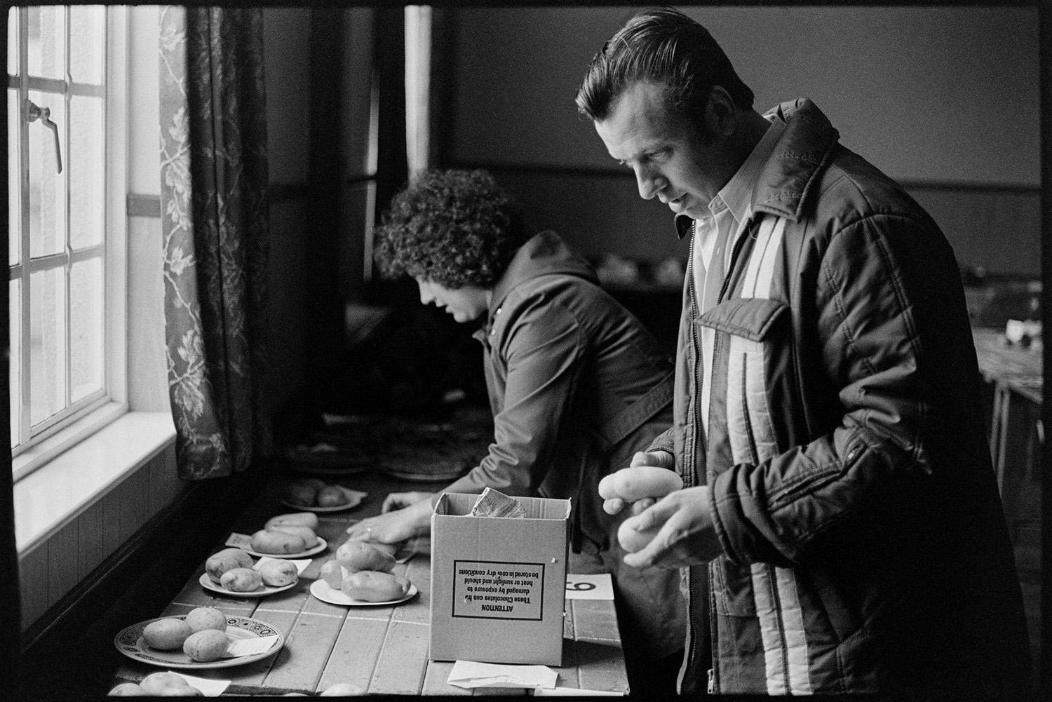 People bringing in entries for flower show and arranging them in village hall. 
[A man and woman arranging potato entries on plates at Dolton Flower Show in Dolton Village Hall.]