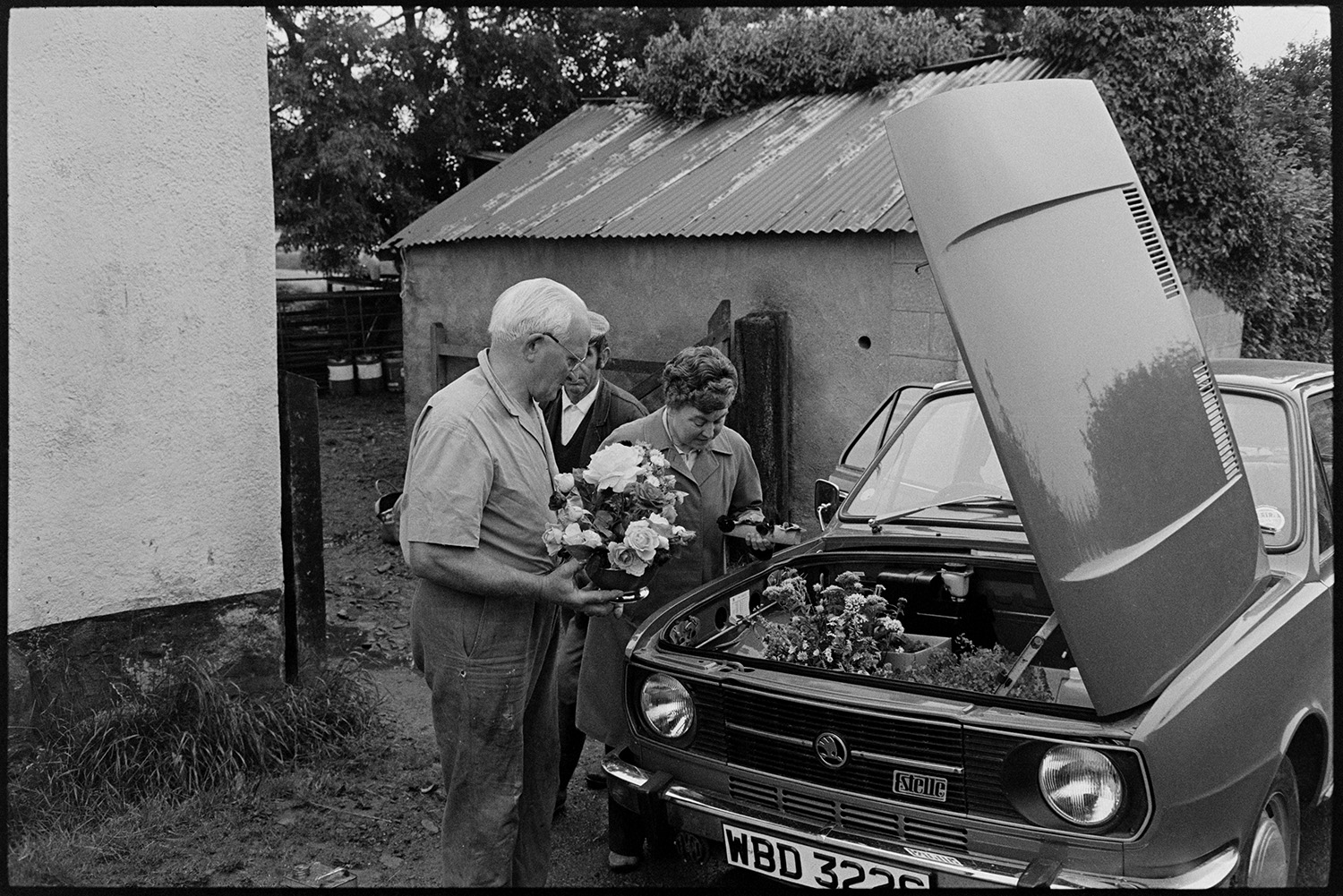 People bringing in entries for flower show and arranging them in village hall. 
[Two men and a woman looking at flower arrangements in a car which they are taking to Dolton Flower Show in Dolton Village Hall. The car storage is under the bonnet of the car.]