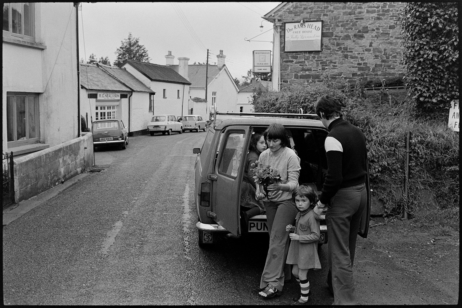 People bringing in entries for flower show and arranging them in village hall. 
[A man, woman and children unloading vases with flower arrangements, from a van parked outside The Rams Head pub in Dolton, which they are taking to Dolton Flower Show in Dolton Village Hall.]