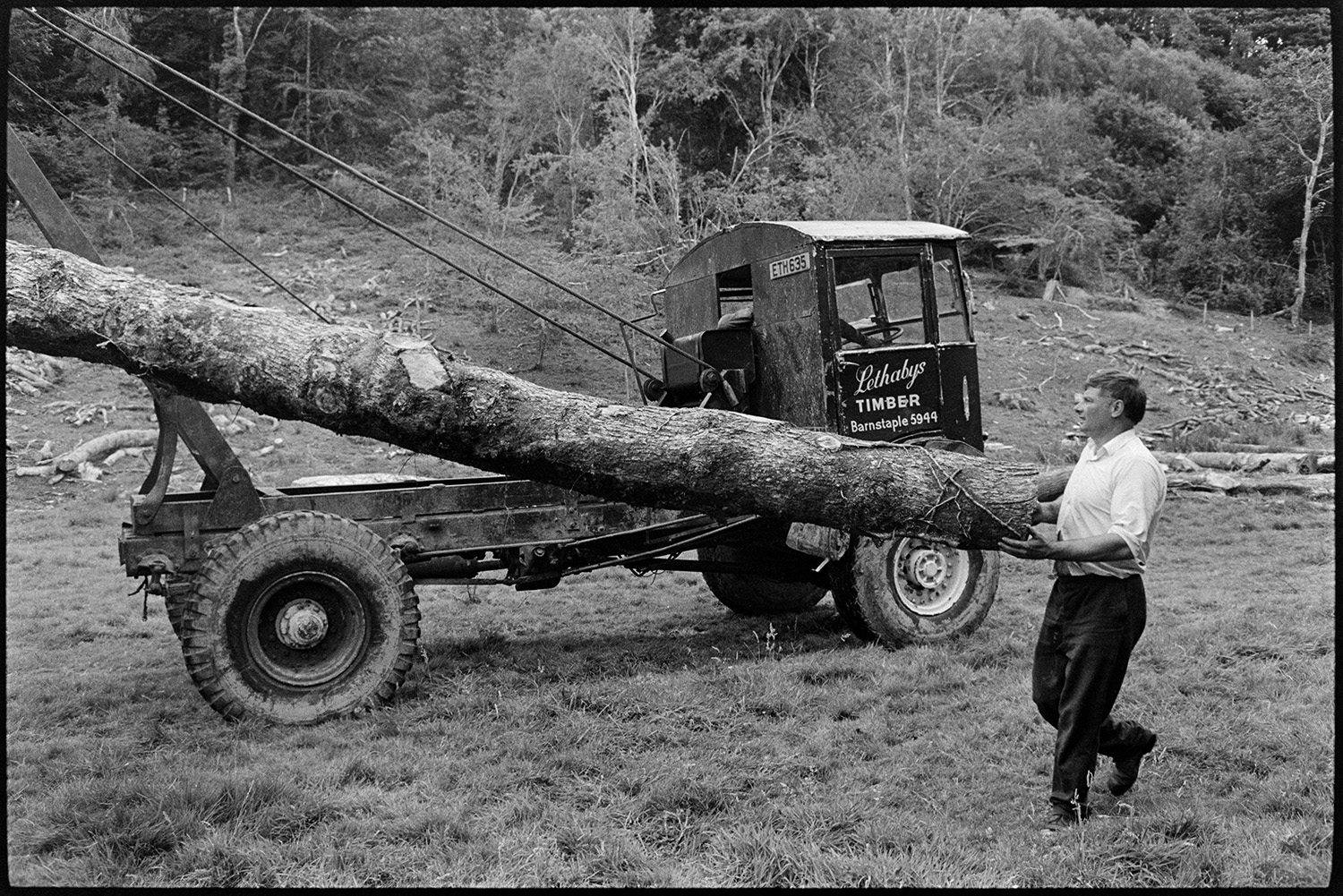 Men sawing up felled timber and loading on to lorry using tractor and winch. 
[A man loading a felled tree trunk onto a lorry using a winch, in a field at Halsdon, Dolton. The winch is from Lethabys timber merchants.]