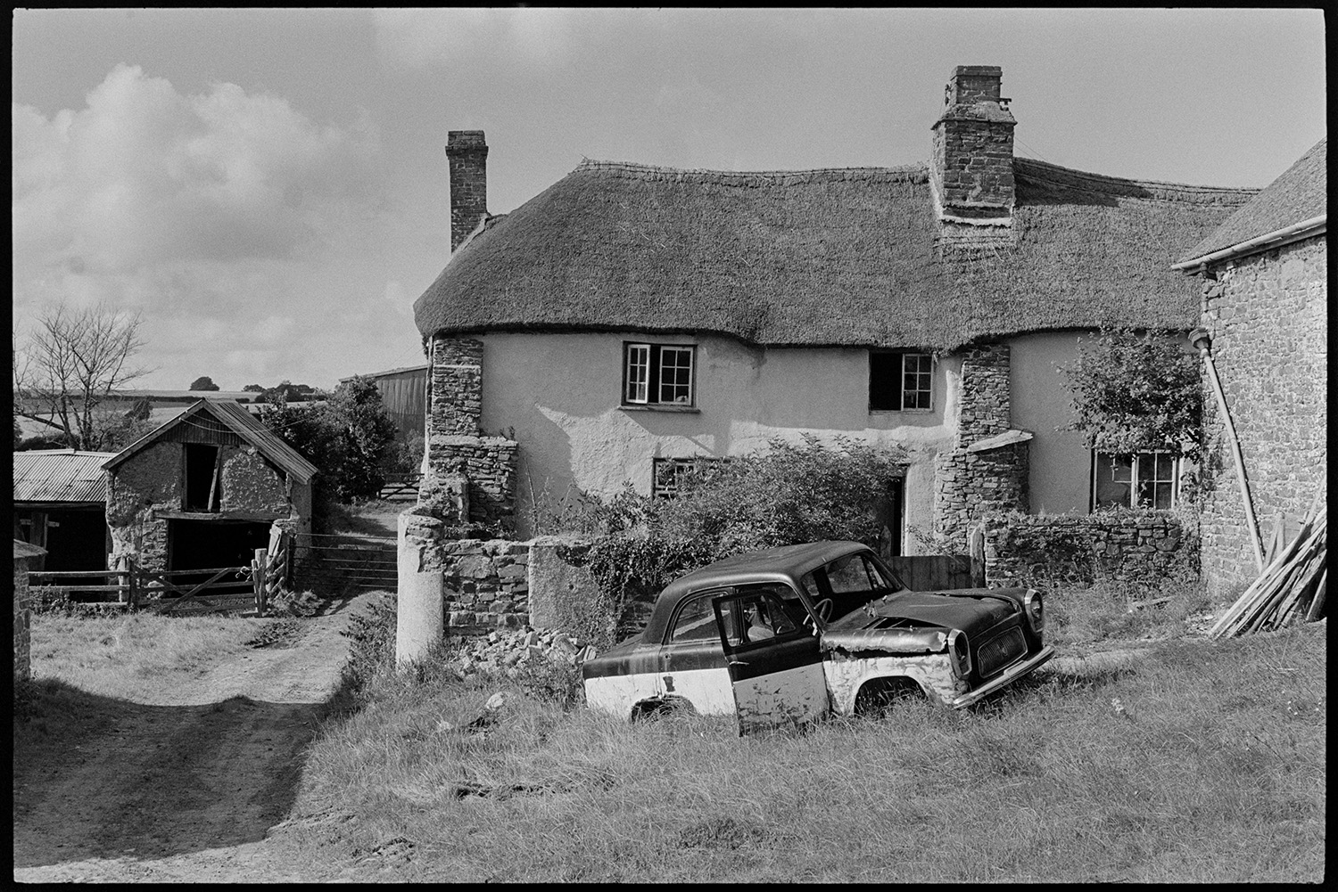 Thatched farmhouse with abandoned car. 
[A thatched farmhouse with out buildings, possibly at Redland, Ashreigney. A abandoned car is parked on the grass in the foreground.]