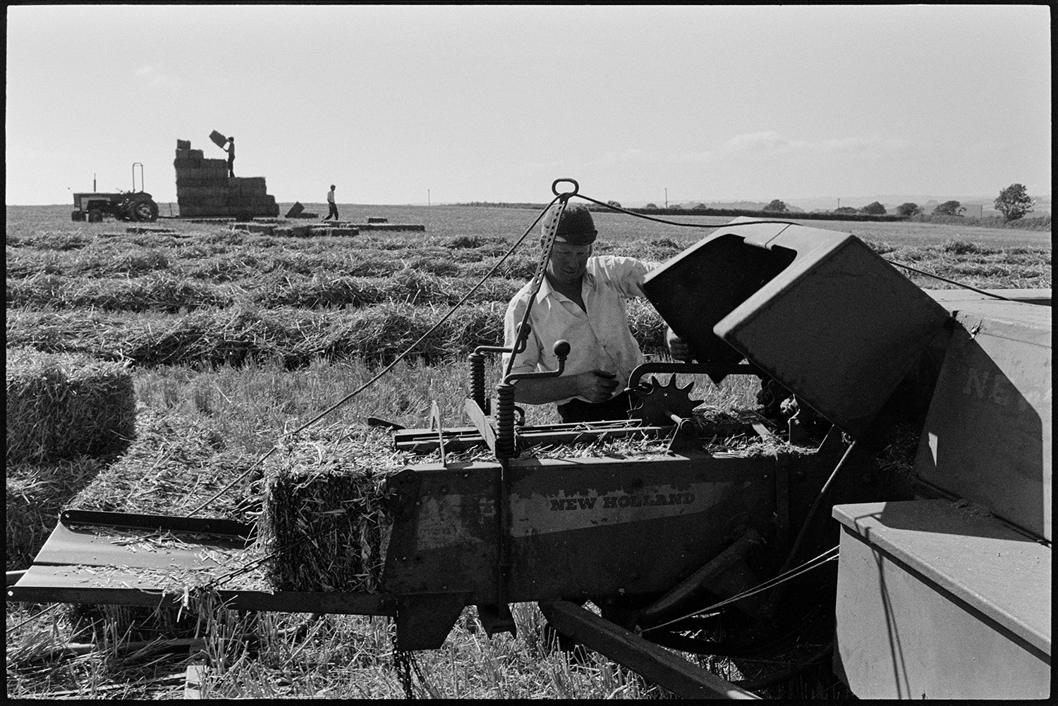 Haymaking, putting hay in barn. 
[A man checking a baler in a field at Dowland. In the background people can be seen loading hay bales onto a tractor and trailer.]