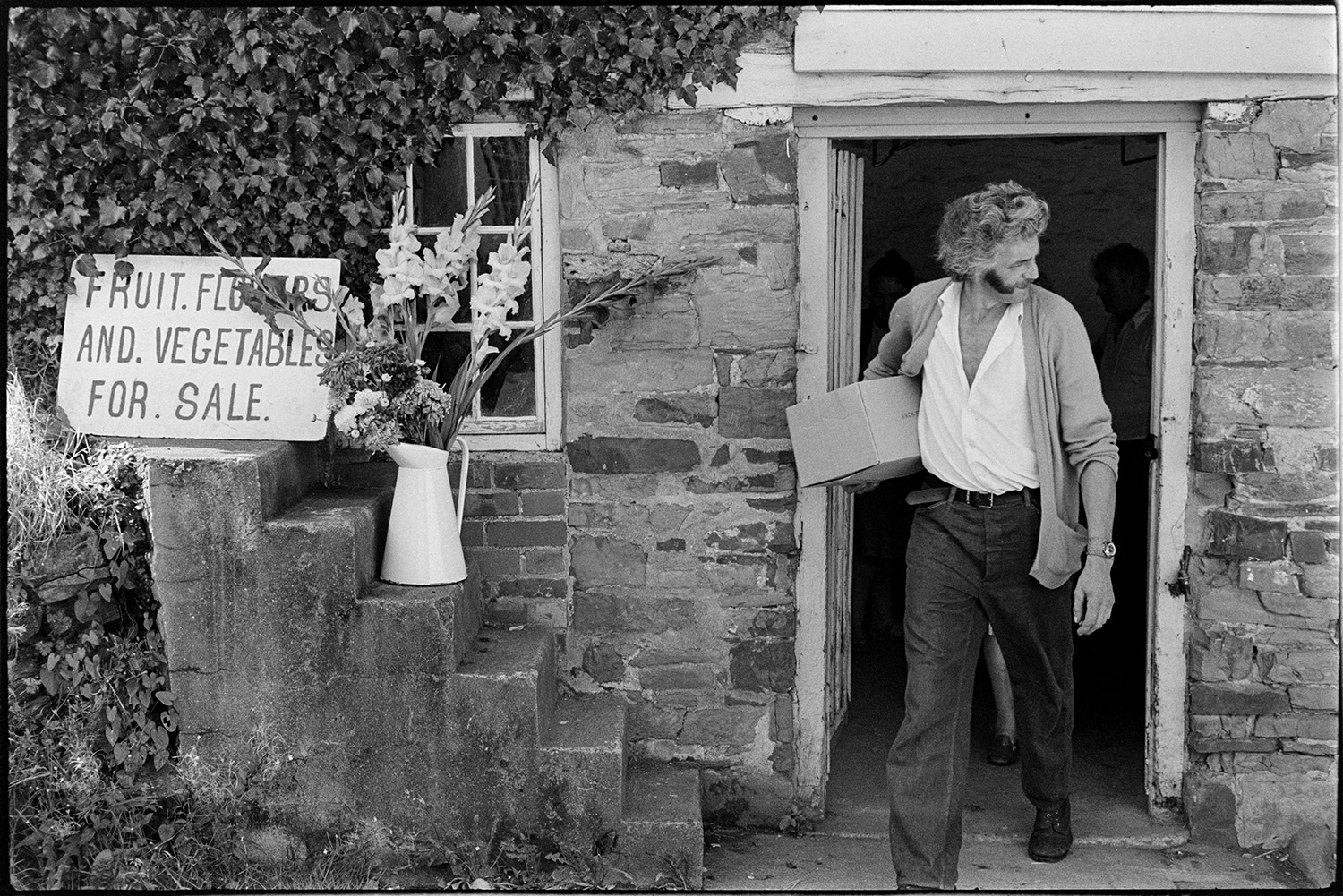 Front of fruit store, putting out flowers. 
[A man bringing a box of fruit or vegetables out of a house to sell on his fruit and vegetable stall in Dolton. A jug with flowers and a sign reading 'Fruit, Flowers and Vegetables For Sale' is placed on steps outside the cottage.]