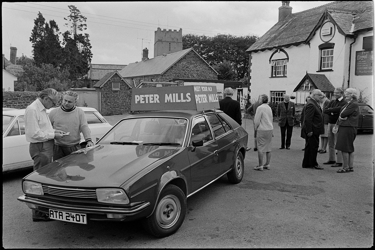 People listening to MP in village, talking to constituent beside his car with placard. 
[Men and women waiting to speak to Peter Mills, MP, outside the Royal Oak pub in Dolton. A car with a placard advertising Peter Mills in parked in the foreground and Dolton Church tower is visible in the background.]