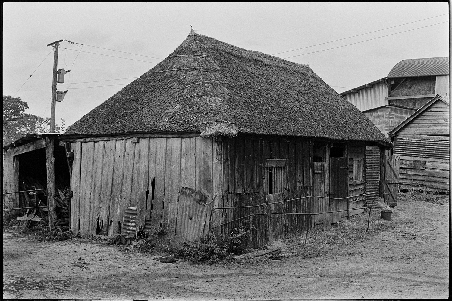 Wooden barn with thatched roof now demolished. 
[A wooden barn with a thatched roof at Leigh Farm, Chulmleigh. Other barns, one with hay bales, and farm buildings can be seen in the background.]