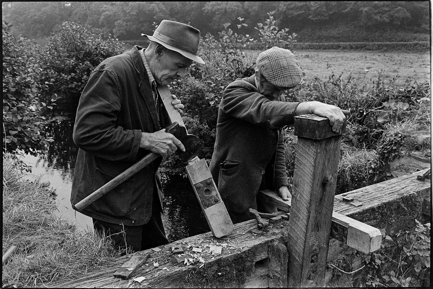 Men repairing wooden sluice gate. 
[Two men repairing wooden sluice gates near Head Mill, Kings Nympton. One of the men is using an axe to shave one of the wooden parts of the sluice gate.]