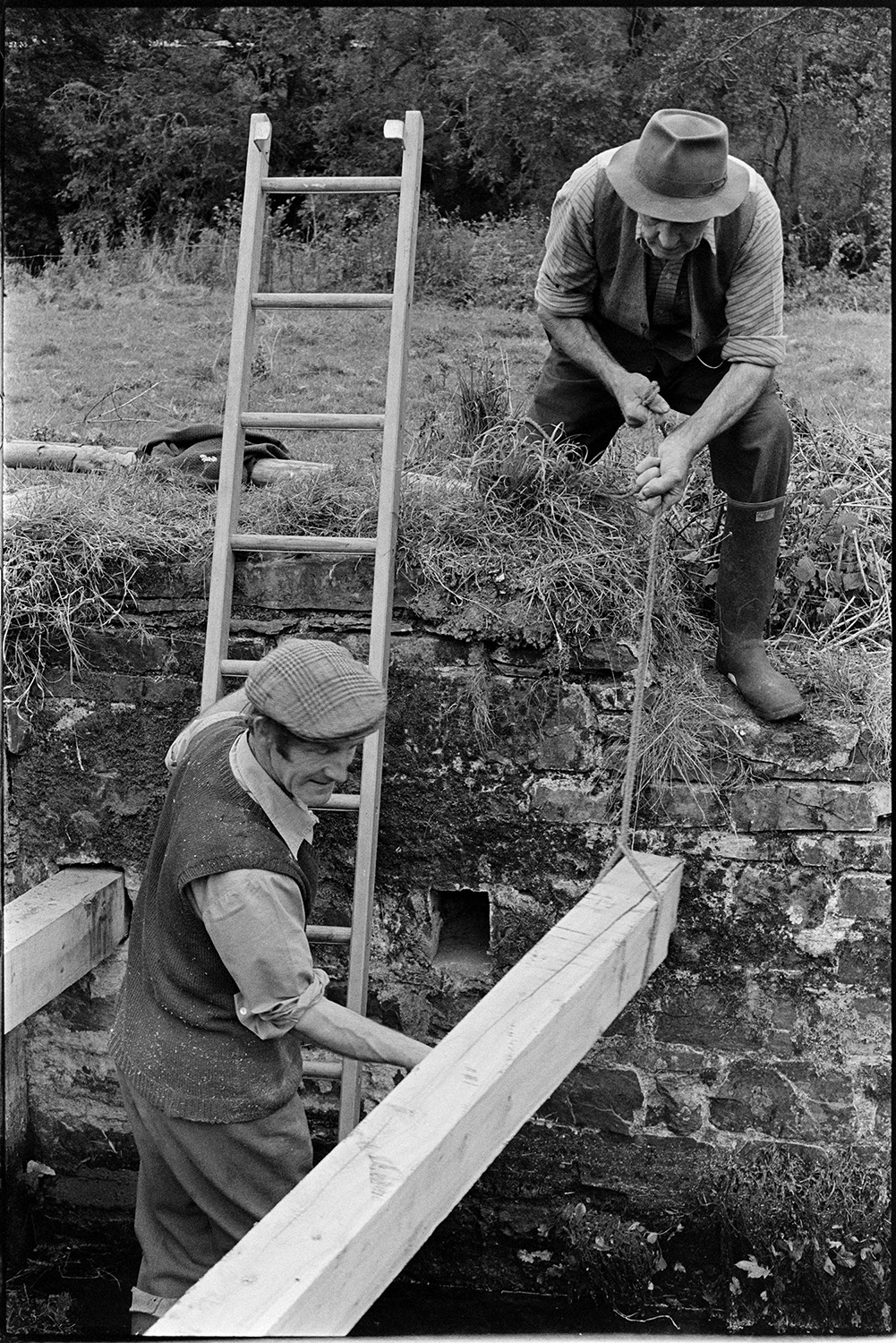 Men repairing wooden sluice gate. 
[Two men repairing a wooden sluice gates near Head Mill, Kings Nympton. One of the men is on a ladder in the sluice and they are lowering a wooden beam into the sluice.]