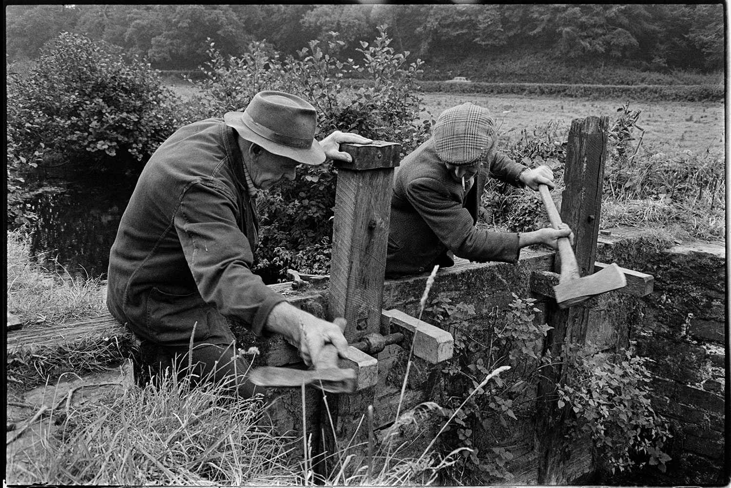 Men repairing sluice gate of mill leat. 
[Two men repairing a wooden sluice gate of the leat running to Head Mill, Kings Nympton. They are using axes.]