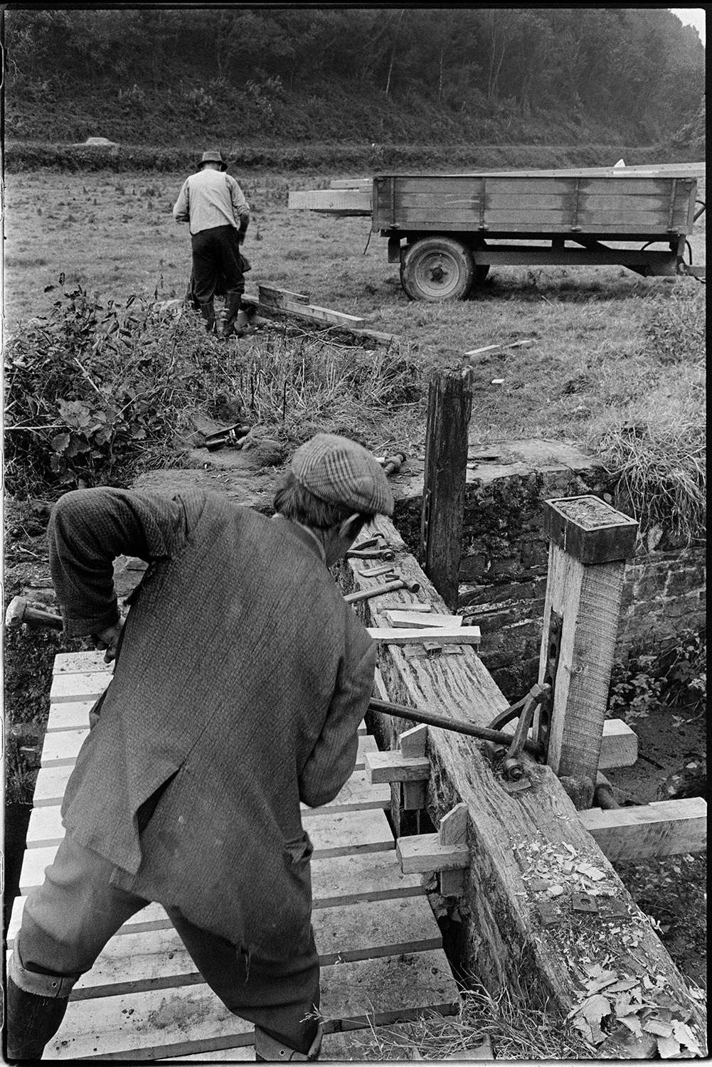 Men repairing sluice gate of mill leat. 
[Two men repairing a wooden sluice gate of the leat running to Head Mill, Kings Nympton. Various tools are resting on a wooden beam of the sluice and a trailer can be seen in the background.]