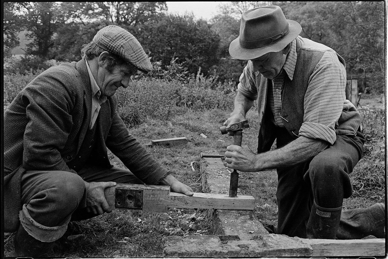 Men repairing sluice gate of mill leat. 
[Two men repairing a wooden sluice gate of the leat running to Head Mill, Kings Nympton.  They are using a hammer and chisel to shape one of the wooden parts of the sluice.]