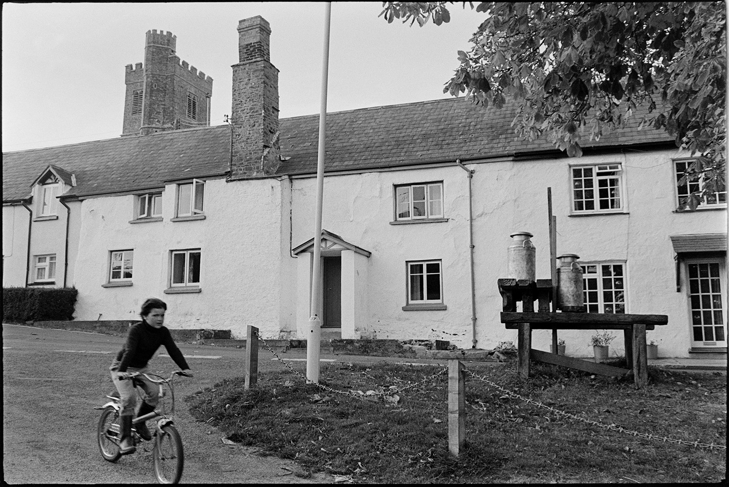Village square, milk churns on stand. 
[A child riding a bicycle past a wooden milk churn stand with milk churns on it waiting to be collected, in Atherington. Terraced houses are visible in the background.]
