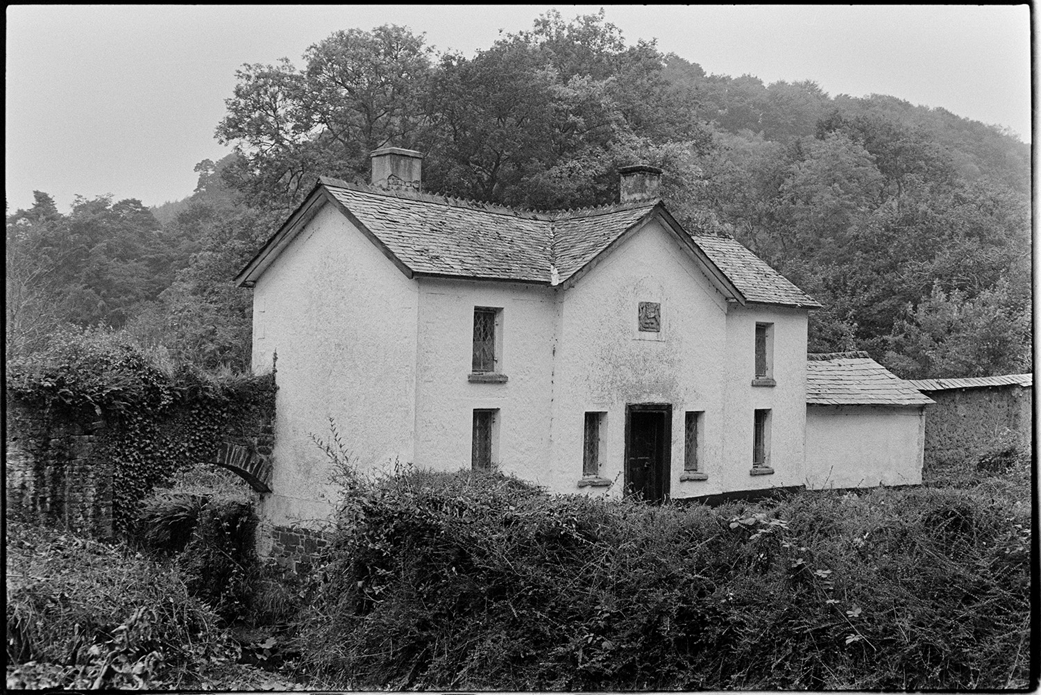 House, lodge at entrance to park. 
[A house with a crest inlaid into the wall, surrounded by trees and a stream at Jewells Bridge, Kings Nympton. A brick and stone archway can be seen over the stream.]