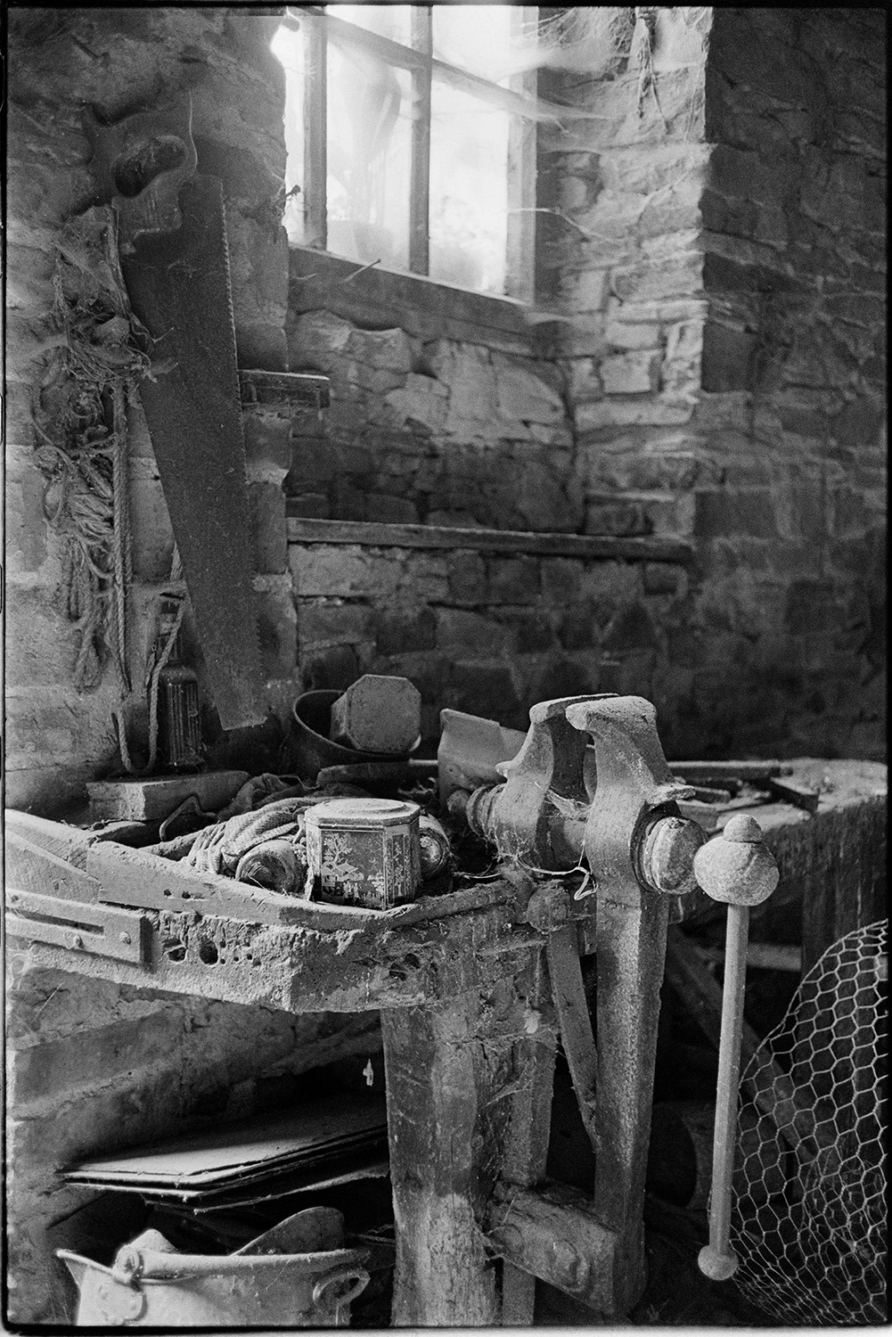 Workbench and vice in old wheelwright's shop. 
[A vice attached to a workbench with various tools in a former wheelwright's workshop by the churchyard path in Beaford. A saw is hung up on the wall behind the workbench.]
