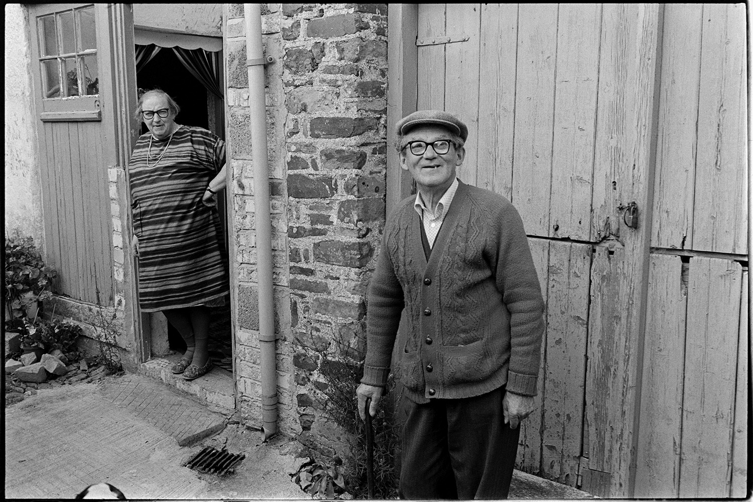 Old couple outside former wheelwright's shop. 
[Mr Bright and Mrs Bright stood by the wooden doorway to a former wheelwright's workshop by the churchyard path in Beaford.]