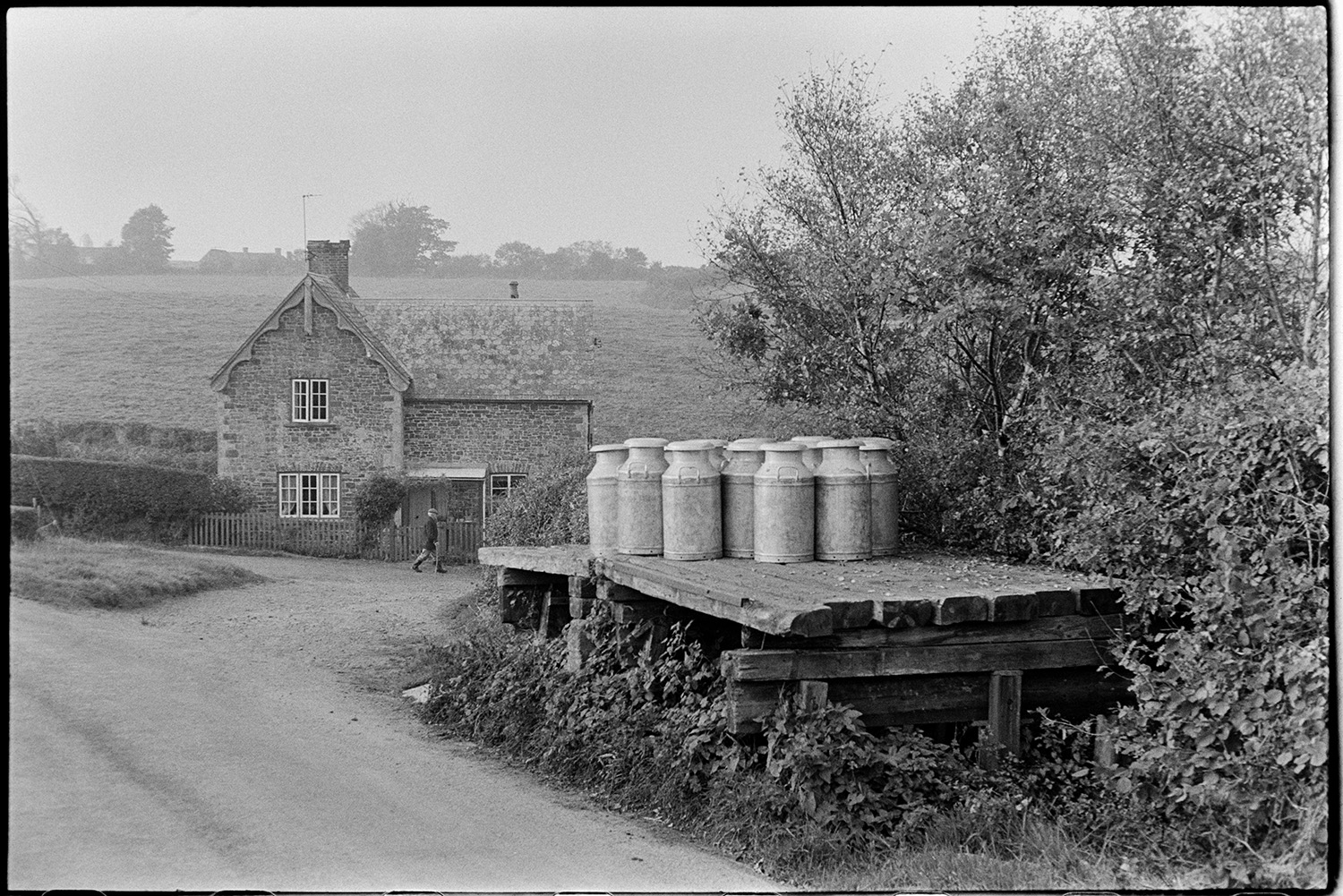 Milk churns on stand. 
[Milk churns on a wooden milk churn stand waiting to be collected at Hallcourt, Petrockstowe. A person is walking past a house in the background.]