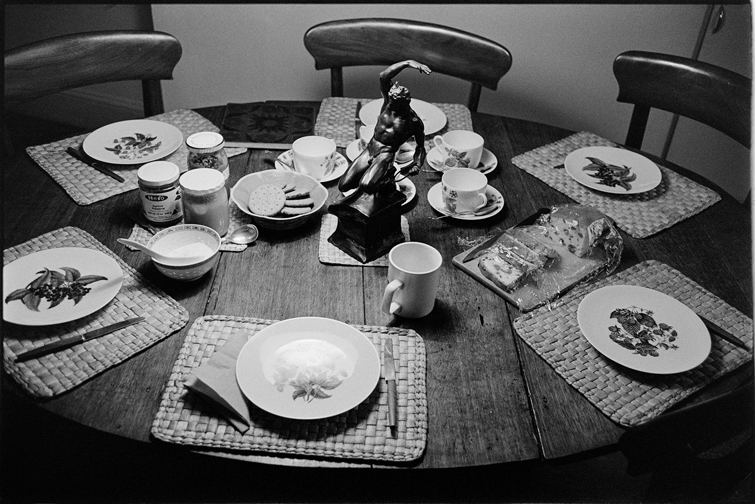Table laid for tea, mugs, plates. 
[A table laid for tea with plates, cups, cake and biscuits in the house of Orea Purnell at Hartland. A statue of a man is in the centre of the table.]